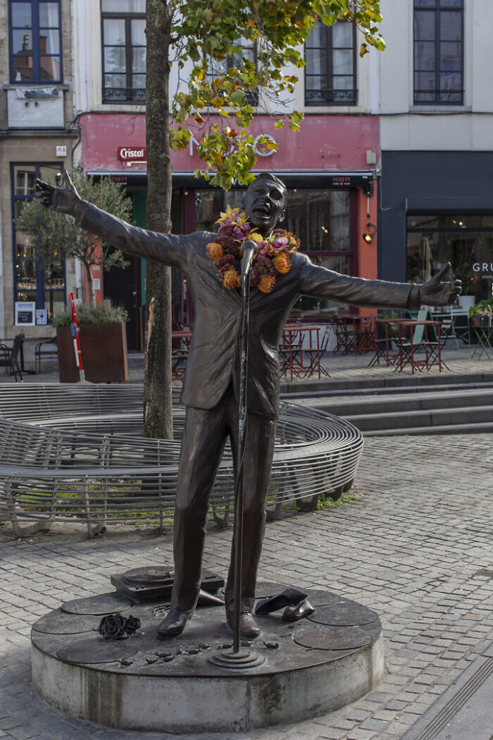 Bronze city statue of a singer decorated with a vibrant flower necklace in an urban plaza setting.