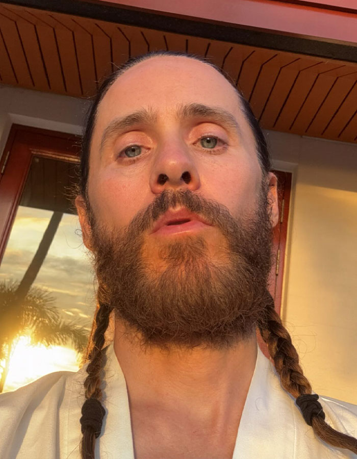 Close-up of Jared Leto with braided hair and beard, facing camera outdoors with golden hour sunlight.