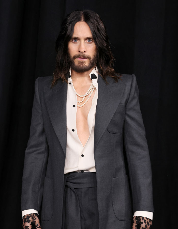 Jared Leto wearing a dark suit and pearl necklace, posing against a black backdrop amid underage claims expos&eacute;.