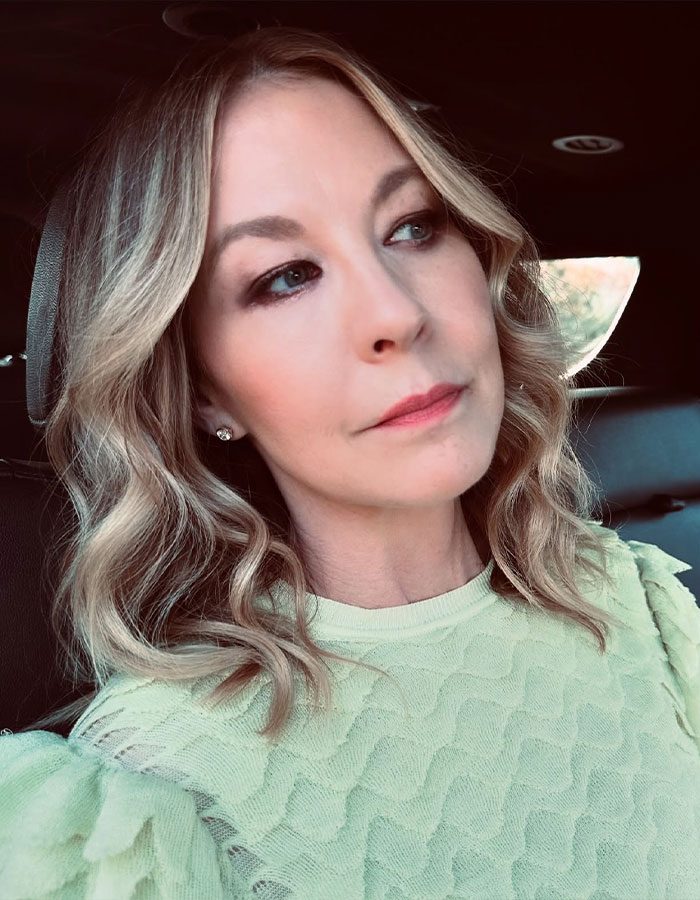 Famous 90s actress aged 53 with youthful look, wavy blonde hair, wearing light green textured top in a car.