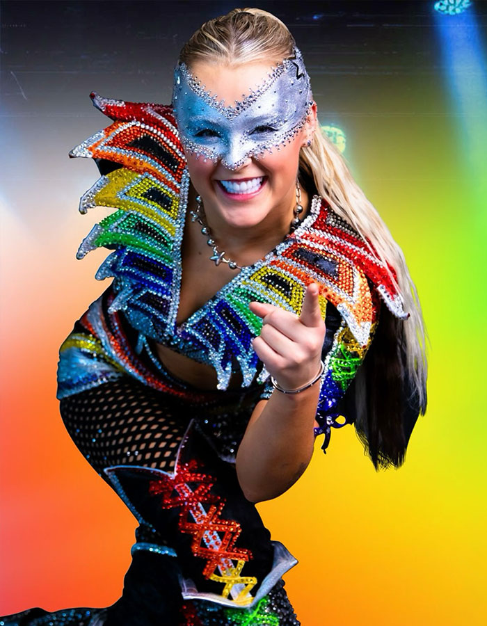 JoJo Siwa wearing a colorful sequin outfit and silver mask, smiling and pointing forward in a vibrant setting. JoJo Siwa wearing a colorful sequin outfit and silver mask, smiling and pointing forward in a vibrant setting.