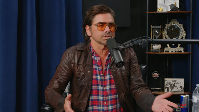 Man in brown leather jacket and tinted glasses speaking into podcast microphone about Hollywood hypocrisy on aging.