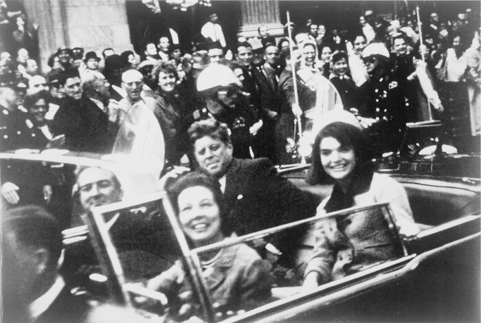 Black and white photo showing famous people riding in an open car surrounded by a crowd in a historic moment.