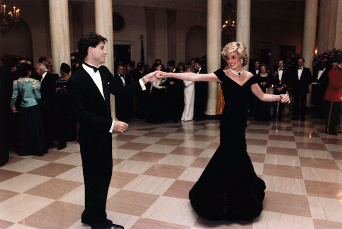 Princess Diana dancing gracefully in an elegant black gown at a formal royal event with guests in the background.