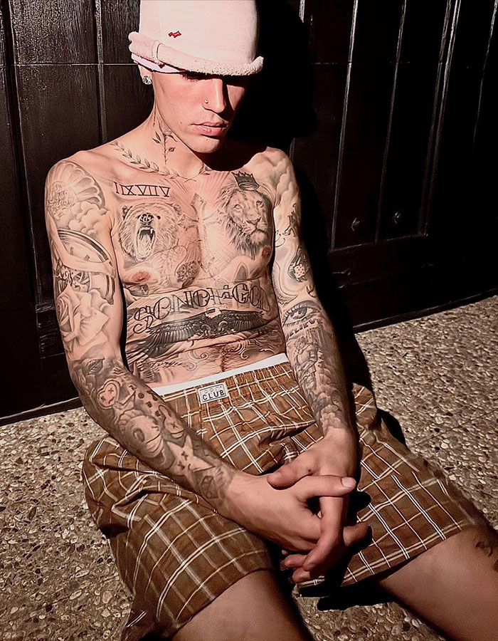 Justin Bieber sitting shirtless in a hat and checkered shorts, showing detailed chest and arm tattoos in dim lighting. Justin Bieber sitting shirtless in a hat and checkered shorts, showing detailed chest and arm tattoos in dim lighting.