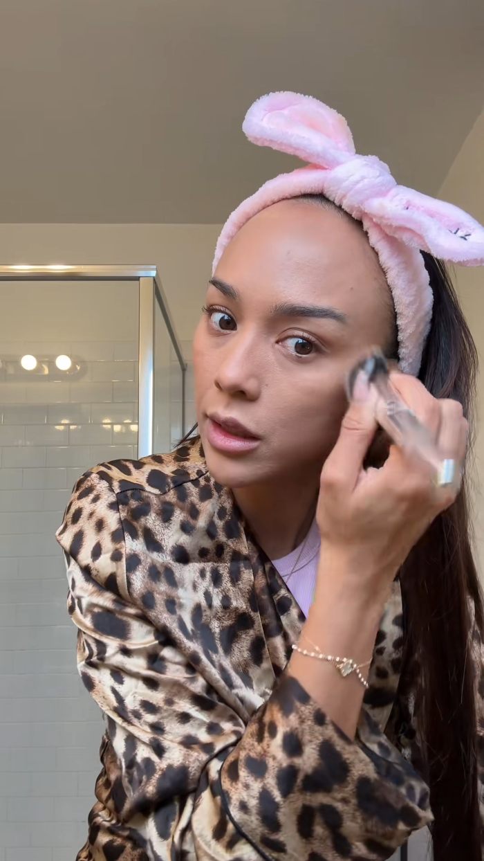 Aussie influencer in the US applying makeup with a pink headband, leopard print robe, discussing cost of living complaints.