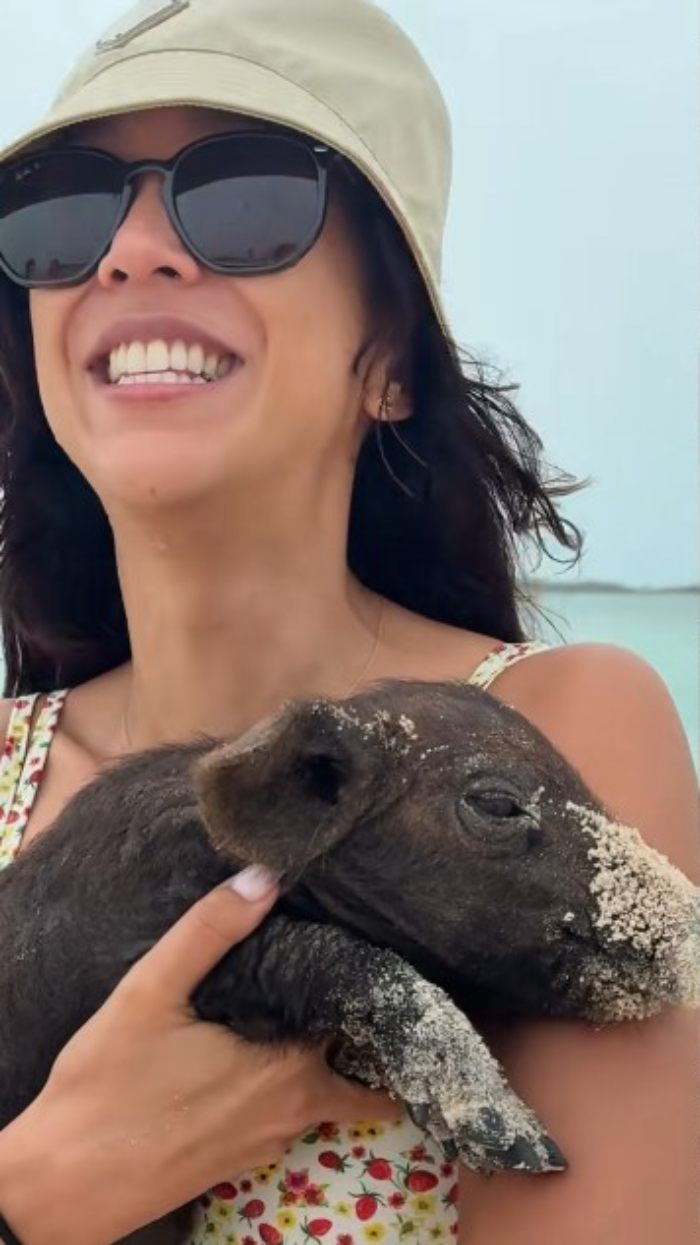 Aussie influencer wearing sunglasses and a hat, smiling and holding a sandy piglet near the beach.