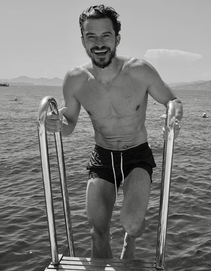 Orlando Bloom smiling while climbing pool ladder, enjoying a swim amid scenic outdoor water and mountain backdrop.