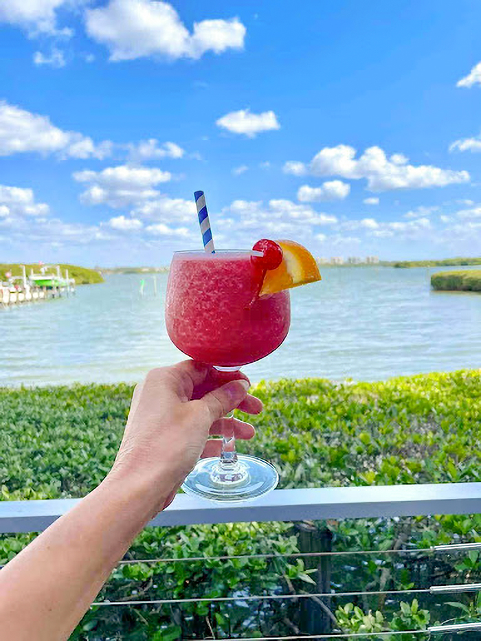 Hand holding a tropical drink with blue straw and fruit garnish overlooking Siesta Key beach and water under blue sky. Hand holding a tropical drink with blue straw and fruit garnish overlooking Siesta Key beach and water under blue sky.