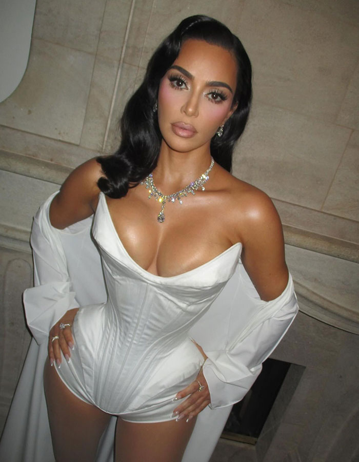 Kim Kardashian posing in a white corset and jewelry in a glamorous setting, with fans discussing her plastic surgeries.