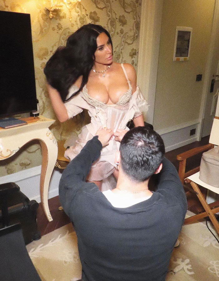 Kim Kardashian posing in lingerie with a stylist adjusting her dress ahead of Bezos pajama party indoors.
