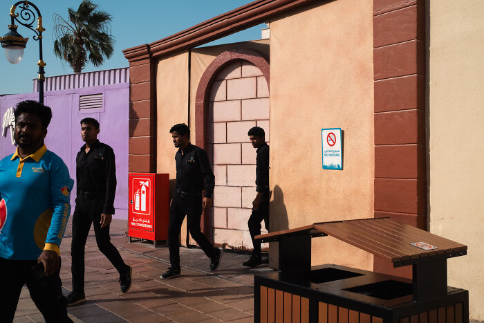 Men walking along a sunlit street near colorful walls and a fire extinguisher box, capturing candid street moments full of emotion.