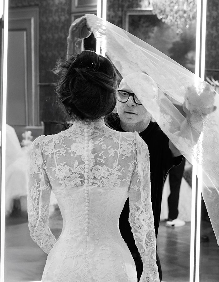 Woman in a detailed lace wedding dress with long sleeves having a veil adjusted, highlighting blue veins on her hands.