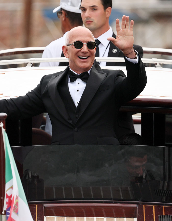 Man in tuxedo wearing sunglasses and waving, with blue veins visible on his hand in a boat setting.