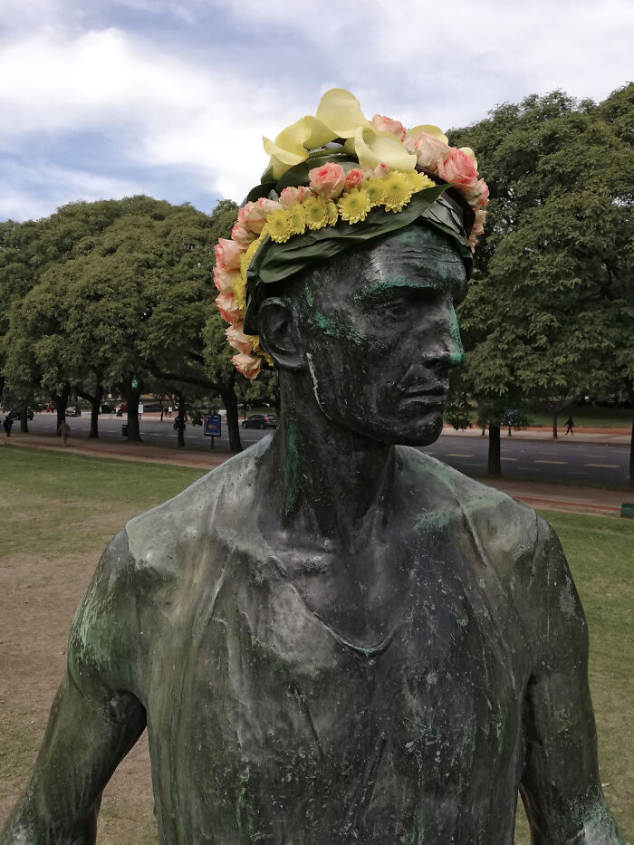Bronze city statue adorned with a colorful floral crown made of yellow and pink flowers in an outdoor park setting.