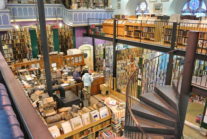 Spiral staircase inside a grandiose cozy library with tall bookshelves and patrons exploring books around wooden desks.