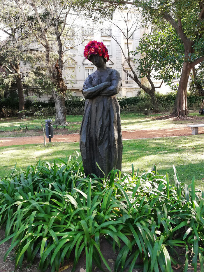 Bronze city statue outdoors adorned with a vibrant red flower crown in a green urban park setting.
