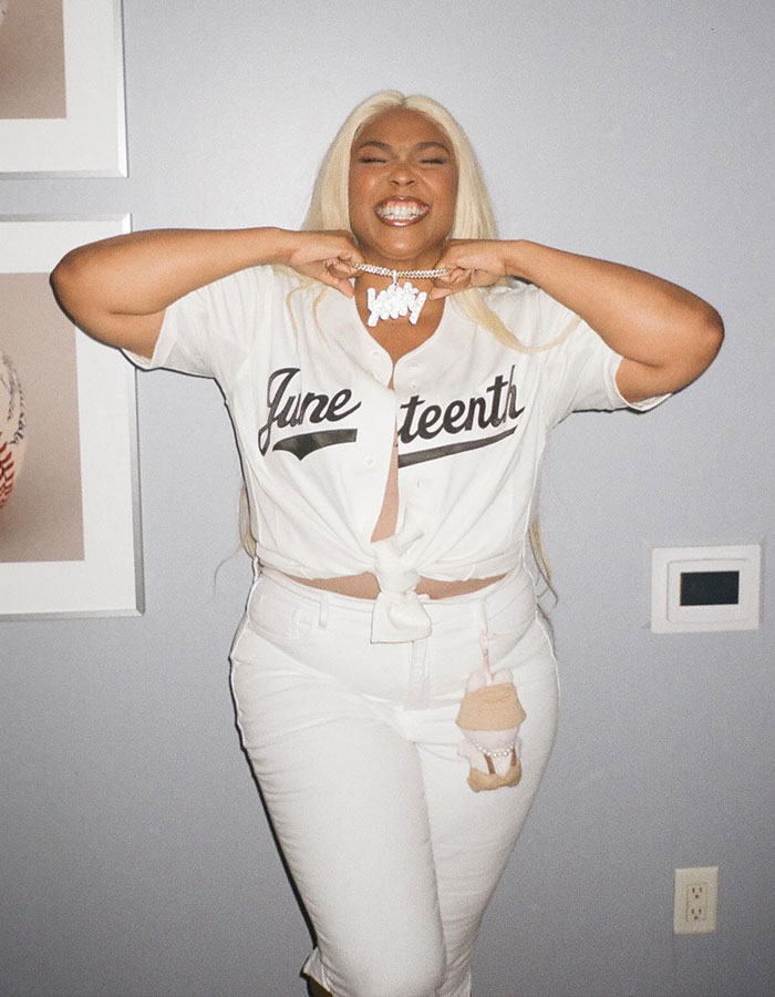 Lizzo smiling and posing indoors wearing a white Juneteenth shirt and white pants, showcasing her body transformation.
