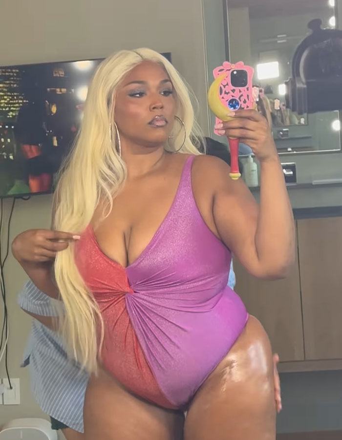 Lizzo showing off mermaid Ozempic body in a pink and red swimsuit, taking a mirror selfie indoors.