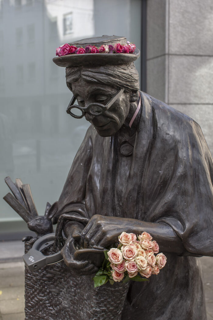 Bronze city statue dressed in flowers on hat and hands, showcasing artistic flower decoration on urban sculpture.