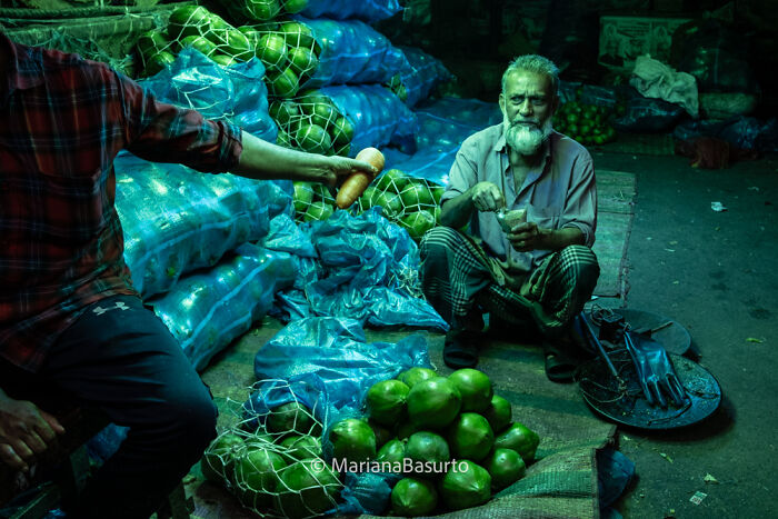Photographer reveals unseen realities in a market scene with an elderly man sitting near green produce and blue bags.
