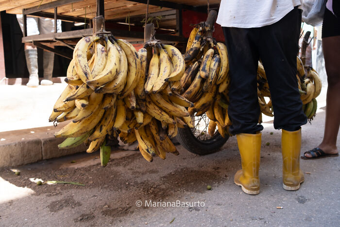 Photographer reveals unseen realities of world with close-up of bananas hanging by street vendor wearing yellow boots.