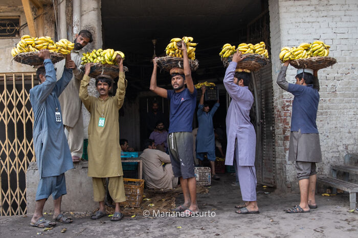 Group of men carrying large baskets of bananas on their heads in a busy market scene, revealing unseen realities of the world.