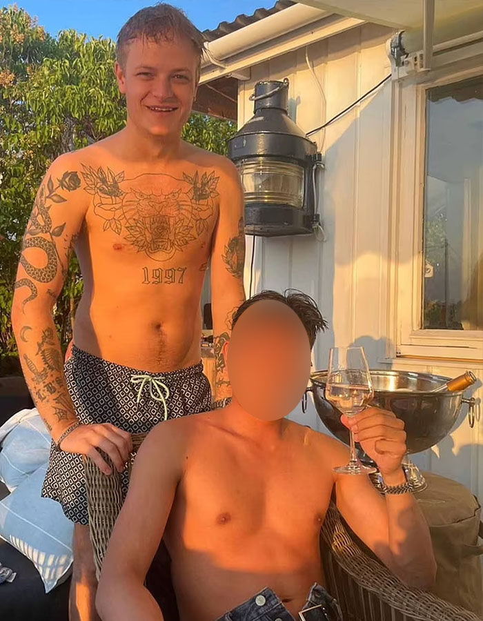 Norwegian Crown Princess&rsquo;s son shirtless outdoors with tattoos, posing with another man holding a wine glass at sunset.