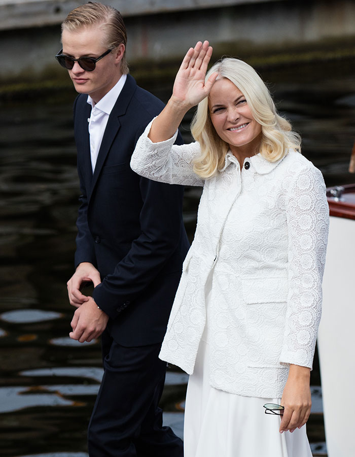 Norwegian Crown Princess&rsquo;s son in dark suit and sunglasses walking by the water while a woman in white waves and smiles.