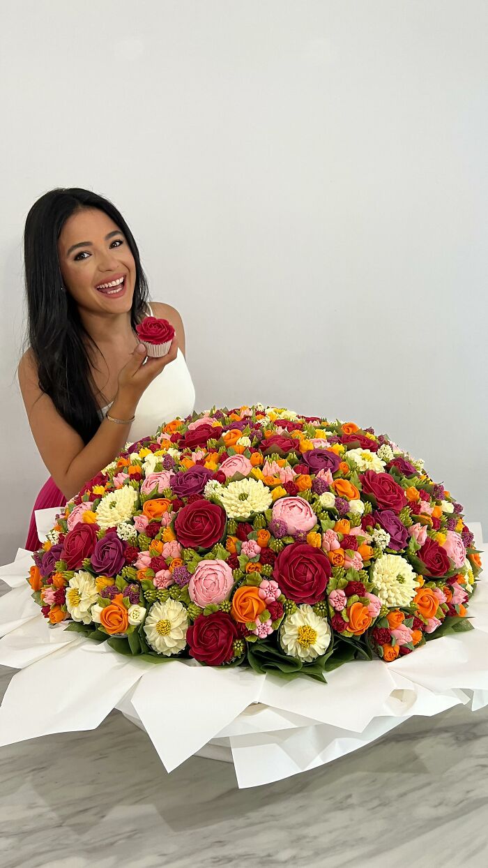 Woman holding a cupcake that looks like a flower, next to a large bouquet of baked goods resembling colorful flower bouquets.