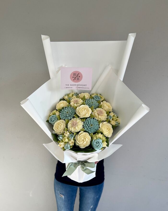 Person holding a bouquet of baked goods that look like flower bouquets in cream, green, and blue tones wrapped in white paper.