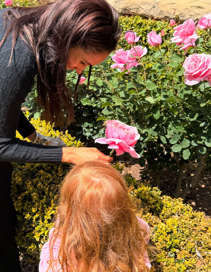 Meghan Markle leaning over garden bushes, holding a pink rose near a young child with red hair in an outdoor setting.