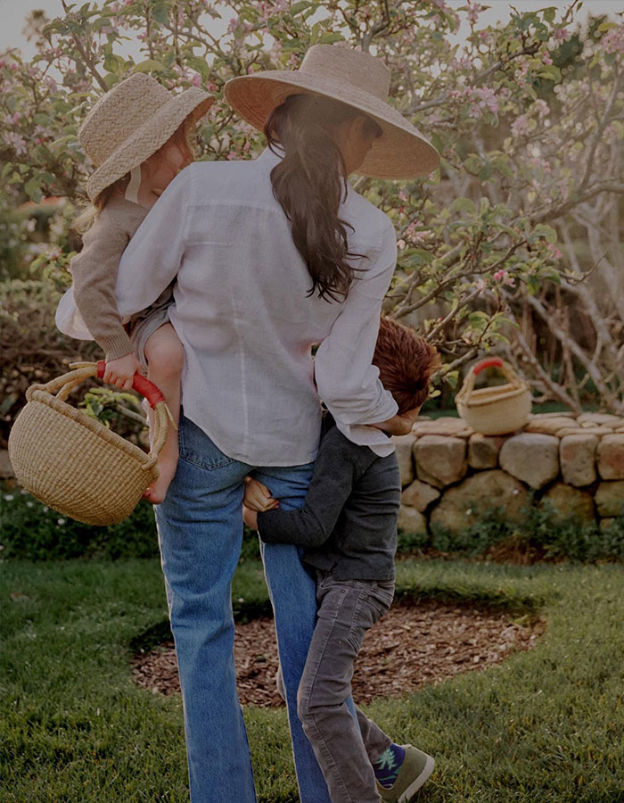 Woman in a hat carrying a child and holding hands with another child in a garden, related to narcissist Meghan Markle focus topic.