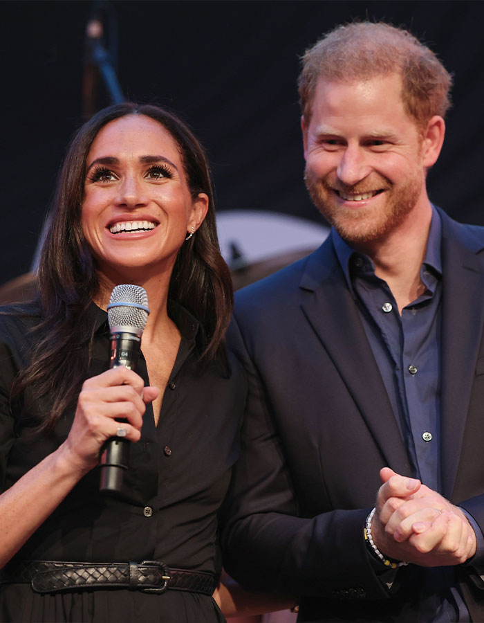 Meghan Markle holding a microphone and smiling beside Prince Harry during a public event with a black background.