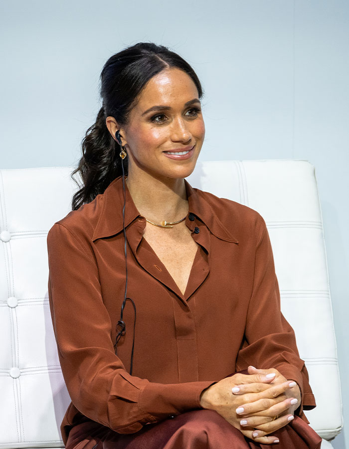 Meghan Markle seated in a brown outfit, wearing an earpiece and smiling during a public speaking event.