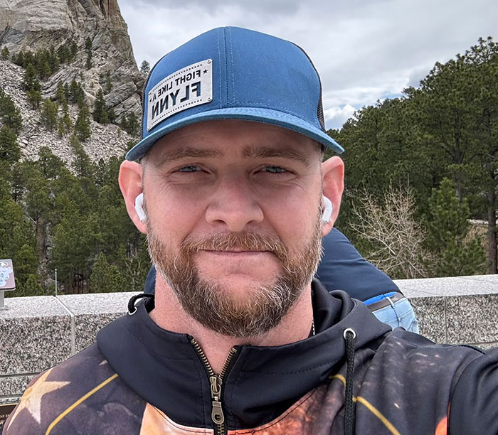 Man wearing blue cap and earbuds posing outdoors near Mount Rushmore, related to Nick Offerman slamming Michael Flynn's son. Man wearing blue cap and earbuds posing outdoors near Mount Rushmore, related to Nick Offerman slamming Michael Flynn's son.