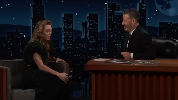 Miley Cyrus discussing her brutal infection with Jimmy Kimmel on a nighttime talk show set with city skyline backdrop.