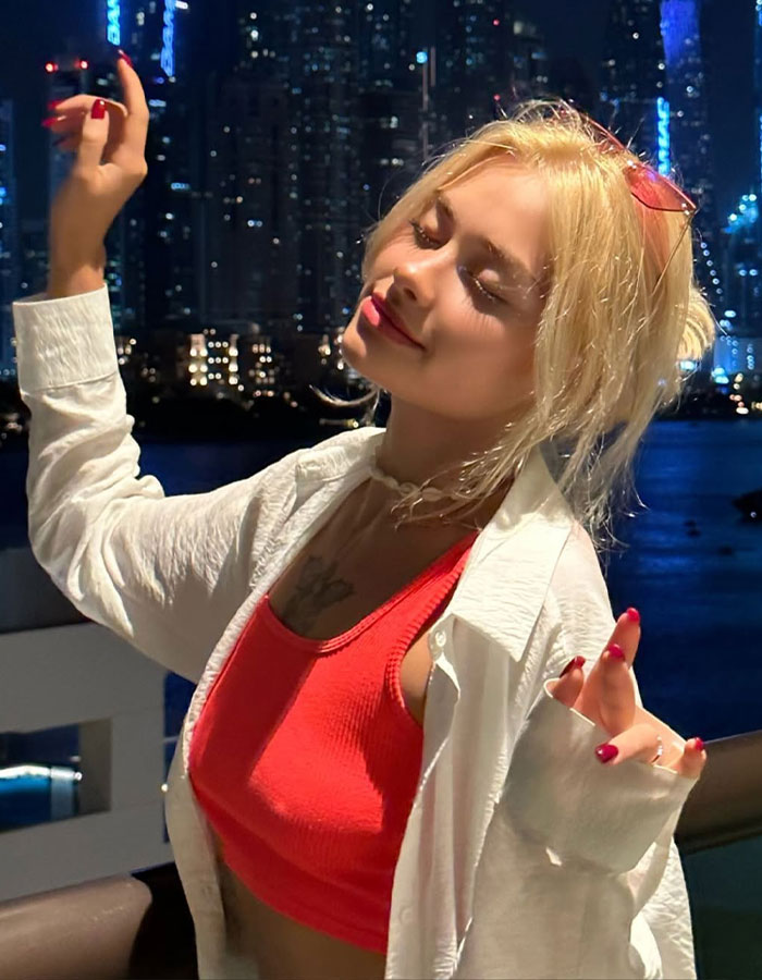 Model in Dubai with closed eyes wearing red top and white shirt at night near city skyline after brutal attack recovery
