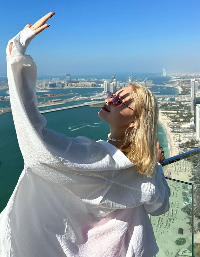 Blonde model wearing sunglasses overlooking Dubai coastline from a high vantage point on a sunny day.
