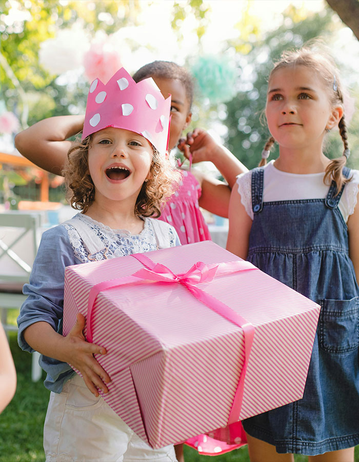Child wearing a pink paper crown holding a large birthday gift with kids in the background at an outdoor party.