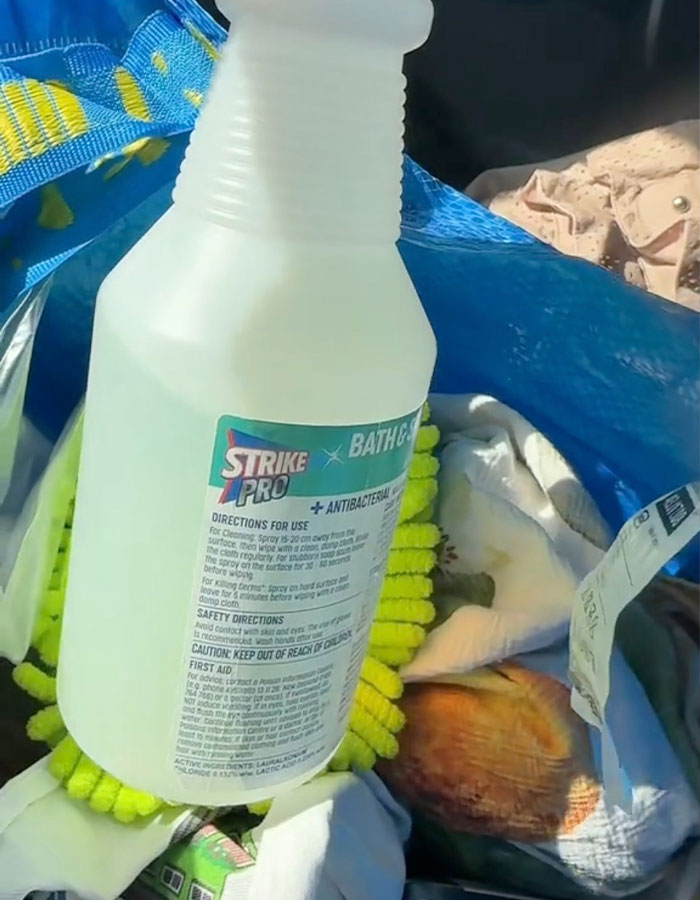 Plastic spray bottle of antibacterial bath and glass cleaner on top of cleaning cloths and rags inside a blue bag.