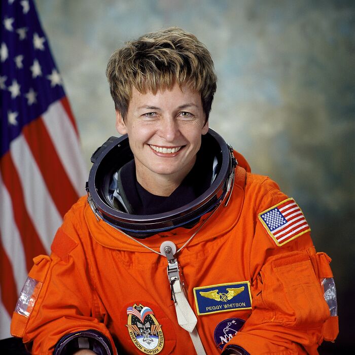 Female astronaut in an orange space suit smiling with American flag background, linked to intriguing and bizarre astronaut facts.