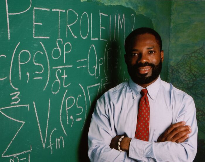 Black scientist in white shirt and red tie standing confidently by chalkboard filled with petroleum formulas, highlighting inventions by people of color