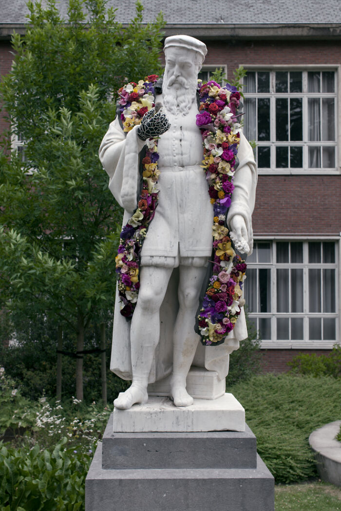 Marble city statue adorned with a vibrant floral arrangement draped over shoulders in an outdoor garden setting.