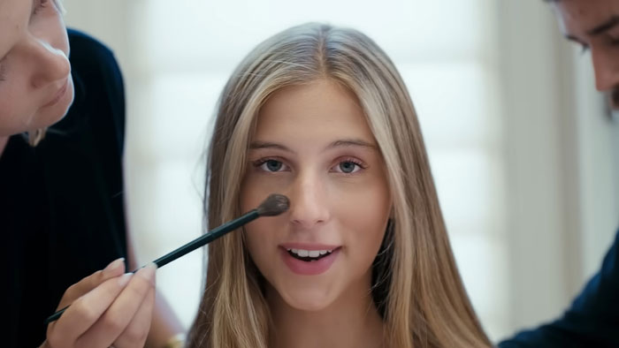Young woman getting makeup applied, representing a nepo grandbaby involved in a song directed by her mom.