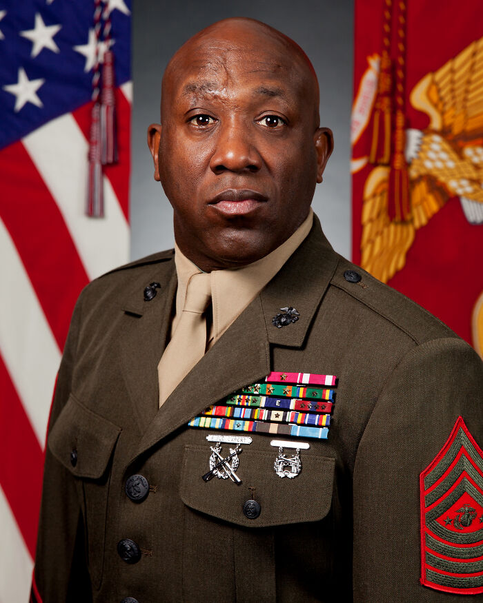 African American military member in uniform with medals standing in front of American and Marine Corps flags.