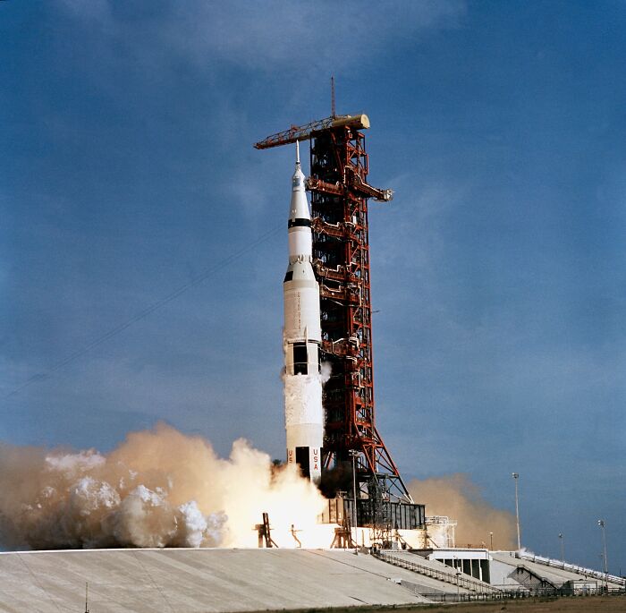 Apollo mission rocket lifting off from launch pad with smoke and flames against a clear blue sky background