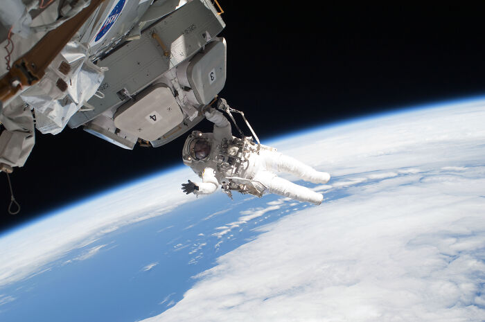 Astronaut performing a spacewalk outside a spacecraft with Earth and clouds visible below in the background.