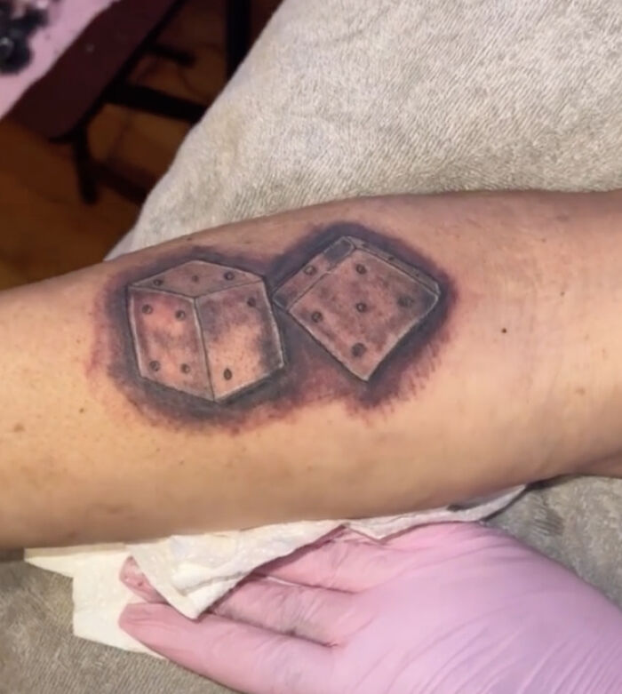 Bizarre tattoo choice showing two shaded dice on a forearm during the tattooing process with a gloved hand nearby.