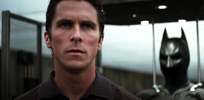Christian Bale portraying Batman, standing in front of a dark armored suit, scene from one of the greatest films.